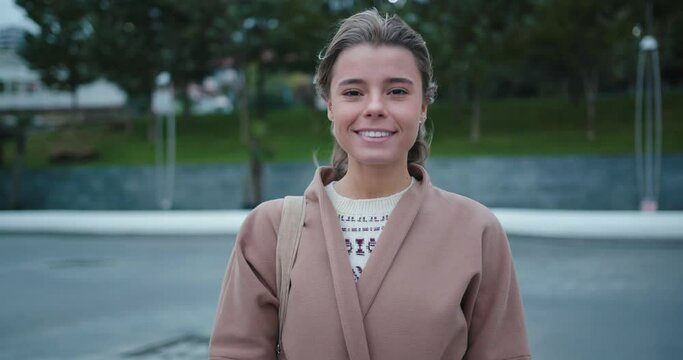 Young blonde long-haired woman with nude make-up and hairstyle in warm coat and shirt stands smiling in deserted windy street slow motion