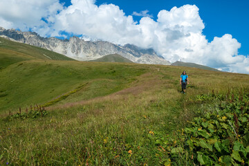 Obraz premium A man hiking along a narrow pathway in high Caucasus mountains in Georgia. There are high glaciers in the back. Thick clouds above the sharp peaks. Lush pastures on the sides. Barren peaks.
