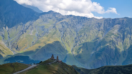 Fototapeta premium A distant view on Gergeti Trinity Church in Stepansminda, Georgia. The church is located on a high Caucasian mountain. Clear and blue sky above the church. In the back there are high mountain chains.