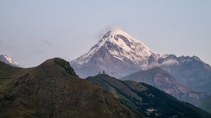 Distant view on Gergeti Trinity Church in Stepansminda, Georgia. The church is located on a high Caucasian mountain. Clear and blue sky above the church. Snow-capped Mount Kazbeg in the back. Daybreak