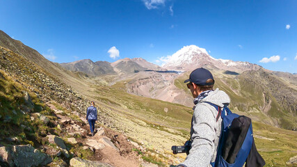 Obraz premium A couple walking on a narrow path towards Mount Kazbeg in Caucasus, Georgia. There slopes are overgrown with green grass. Massive Gergeti Glacier under the sharp peak. Cloudless sky. Exploring.
