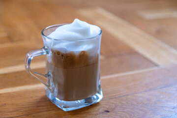 a glass cup of coffee topped with white milk foam on wooden surface