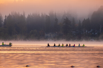 Silhouette of a crew rowing on a lake as the rising sun casts a golden glow over the water and...