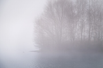 Thick fog over lake in early autumn morning, bare trees silhouetted in the haze