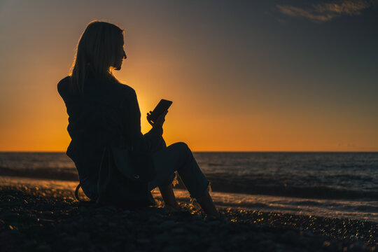 Young woman is sitting on a pebble beach by the sea with a mobile phone in her hands at sunset, a photo against the sun. Concept of happiness, travel, lifestyle