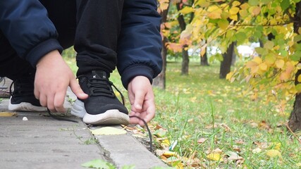 Closeup of unrecognizable sport boy tying sports shoes during evening run outdoors. a schoolboy teenager in a warm jacket walks in an autumn park with colorful foliage. Outdoor fun by any weather
