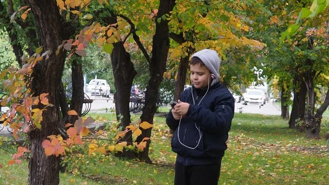 a schoolboy teenager in a warm jacket and a hood walks in an autumn park colorful foliage. with headphones, listening to music or talking on the phone. happy child playing dreams outdoors in autumn