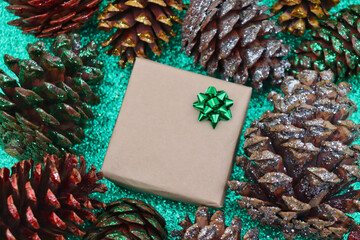 Present wrapped in brown paper with green bow surrounded by silver, green, red, and gold glitter pine cones on green glitter background.