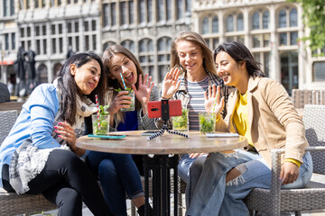 Four multi ethnic female friends, tourists or students sitting at a cafe terrace in the city center using mobile phone for a video call. High quality photo