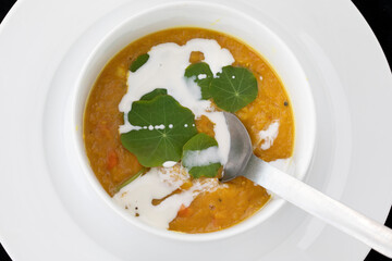 CLOSE UP WHITE BOWL OF NUTRITIOUS CARROT & LENTIL SOUP WITH NASTURTIUM LEAF GARNISH TOPPED WITH  SPLASH OF OAT CREAM ON PLATE ,SPOON