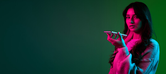 Studio shot of young adorable cute girl, student in casual style clothes posing isolated on dark green studio backgroud in pink neon light. - obrazy, fototapety, plakaty