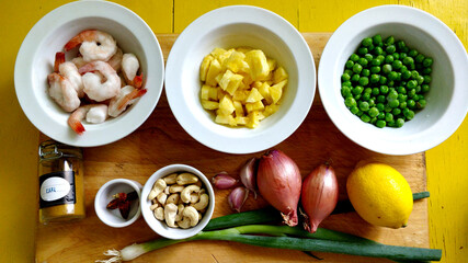 Ingredients prepared for cooking. It contains shrimp, pineapple, peas, onion, lemon, curry, garlic, cashew, and chili. They stand on chopping board. Some ingredients are put into white bowl. The table