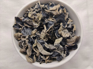 Detail of dried black Jew's ear (wood ear) mushrooms in a white bowl sitting on the white table cloth. The mushroom is type of black Chinese wood ear.