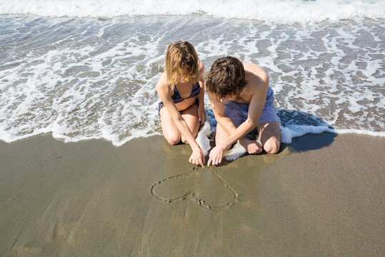 young couple in love, a woman and a man, draw a heart on the sand on the sea coast. Happy holidays, first love, happy relationships, summer vacation, sunny bathrooms