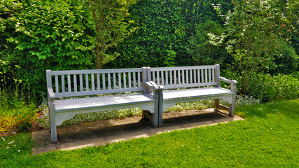 White double bench with hedgerow in the background. There is grass all over the picture and seat looks very relaxing.