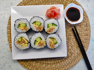 Nicely decorated plate with six pieces of sushi, few pieces of ping pickled ginger, soy sauce and pair of chopsticks. The sushi is filled with cucumber, tuna, and egg in it.