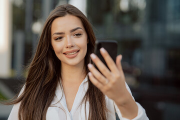 Fototapeta premium A pretty girl stands leaning against a railing in front of a modern glass office building, businesswoman dressed in a white shirt holds phone in front of her taking pictures selfies taking video call