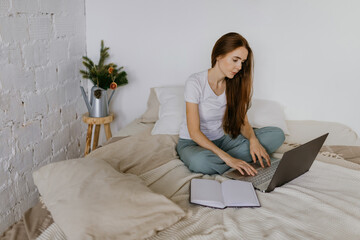 A beautiful woman working remotely at home takes notes on a computer while sitting on a bed