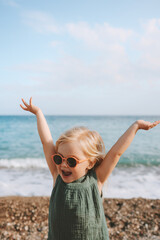 Happy child girl playing on beach emotional toddler in sunglasses 3 years old kid raised hands...