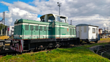 Fototapeta na wymiar Green locomotive parked close to the main train station with small white railway carriage being connected to it. The machine is a diesel one.