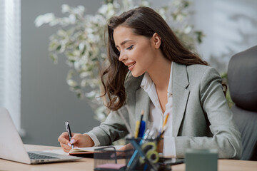 Smiling attractive pretty brunette woman in classic makeup wearing shirt jacket, sitting at desk in a company in front of a laptop, a secretary writes in a calendar appointments, important information
