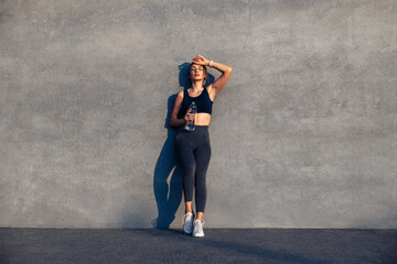 Tired sporty woman holding bottle of water after morning workout, woman standing on city street in front of gray wall