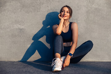 Beautiful fitness woman, in sportswear, happy woman sitting on the floor against the background of a gray city wall