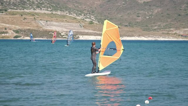 Beginner windsurfer practicing balancing and sailing on a board with an orange sail during a windsurfing lesson at a coastal resort on a sunny summer day