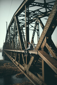 Man in blue denim jacket sitting on rusty brown iron bridge