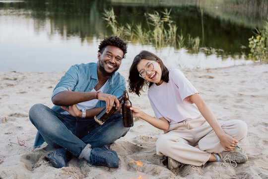 Couple of friends are sitting next to each other on beach by lake smiling laughing woman of Korean Asian beauty is holding bottle of beer in hand is leaning towards man tapping each other cheers - Powered by Adobe