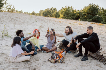 Longtime friends meet at the lake on the beach by the fire, sit on the sand on wooden logs drink beer in bottles raise them toast cheers, laugh, talk, tell stories, students of different nationalities