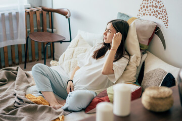 Obraz premium Relaxing pregnant young woman is enjoying sitting on the bed in the bedroom and touching her big belly. Happy motherhood, health care. Gentle caring woman alone at home.