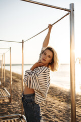 Attractive caucasian young girl cute posing for photo by sea holding one hand to pipe. Blonde with bob haircut, wearing striped blouse and dark jeans. Rest and recovery concept.