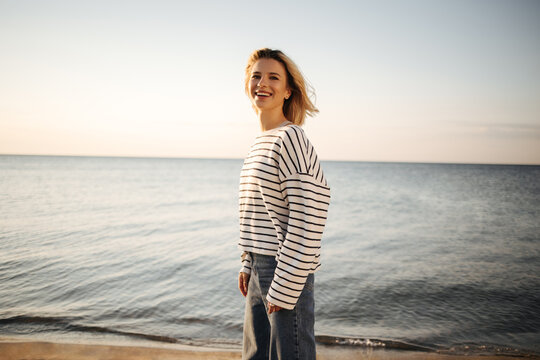 Happy pretty caucasian young woman stands on seashore smiling cute with teeth. Sophisticated blonde dressed in striped black and white blouse and dark jeans. Rest time concept.