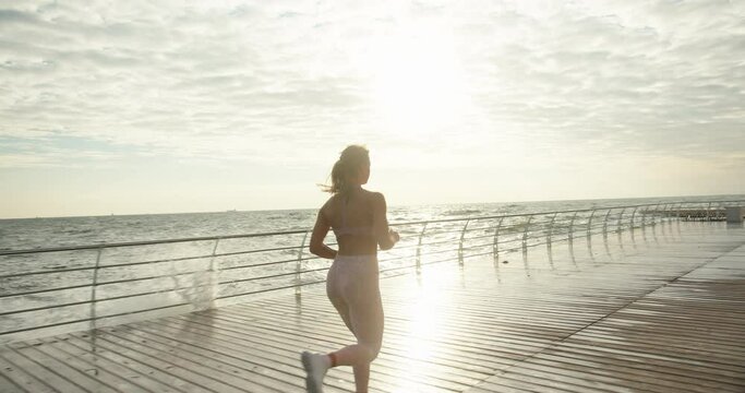Young woman with long blonde ponytail wearing sportswear jogs along waterfront past stormy sea waves on nasty day slow motion