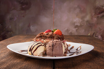 Chocolate cake, a tasty slice of chocolate cake with strawberries and ice cream balls on a white plate on rustic wood, abstract background, selective focus.