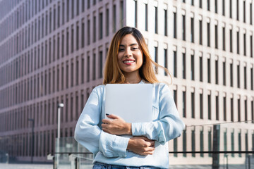 Fototapeta premium Lovely brunette entrepreneur smiling outside surrounded by buildings while posing with a laptop. Business concept