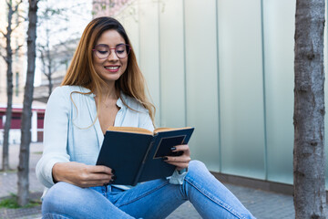 Fototapeta premium Positive optimistic brunette woman with glasses smiling while reading book outdoors. Student concept