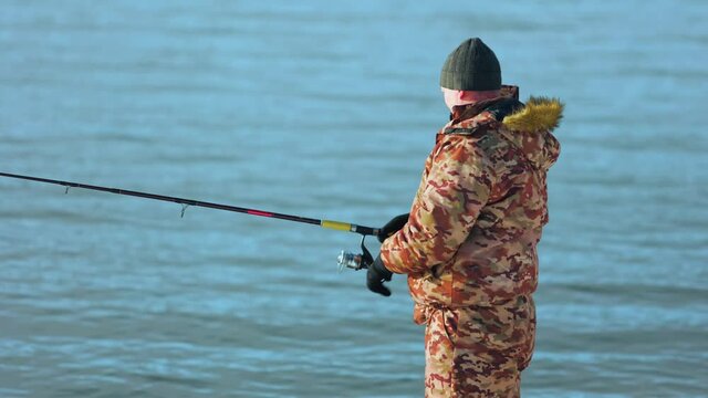 A fisherman in winter gear is fishing on a spinning rod. A man stands against the background of the river. He turns the reel and pulls the fishing line with the bait to himself. Winter fishing.