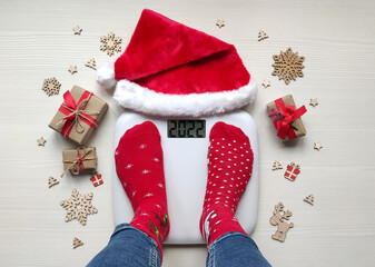 Female feet standing on electronic scales for weight control in funny red Christmas socks on white wooden background.