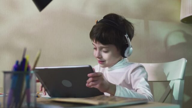 Curious Child Little school Boy Sitting At The Table Listening To Music Using Digital Tablet Technology Device Watching Video Playing Games Internet Education Concept