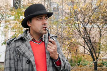 Portrait of a man in a hat and coat smoking a smoking tube outdoors in a park.