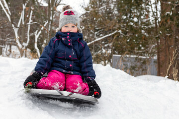 Fototapeta na wymiar Little girl tobogganing downhill, having fun. Small happy child sledging in snow outdoors in winter. Little girl in hat and jacket sitting on sledge in cold weather.