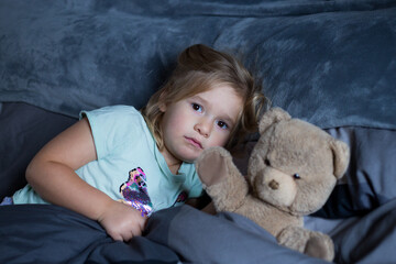 High angle selective focus horizontal view of pretty toddler girl with upset expression cowering in bed next to her teddy bear
