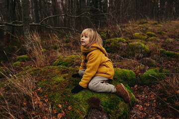 Blonde Child in Yellow Coat Climbing Mossy Stones in Autumn Forest