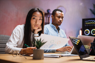 Asian woman and african man ain formal clothes working late at office with documents and gadgets. Focus on pretty Korean lady looking at camera while working with papers