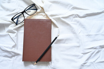 Stationery on a background of white crumpled fabric. Flat lay. Composition with a place under the inscriptions. Layout from a closed book, pencil and glasses