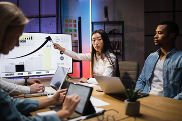 Four multi ethnic economists sitting at office with various gadgets and analysing financial growth in worldwide economy. Male and female partners having business meeting during evening time.