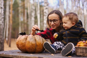 Young woman and her baby son in autumn park, boy playing with helloween pumpkin and eating pumpkin bun