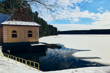 Zimowy spacer wokół jeziora. Zamarznięta tafla pokryta jest białym śniegiem. Na brzegu domek, budowla hydrologiczna. - obrazy, fototapety, plakaty